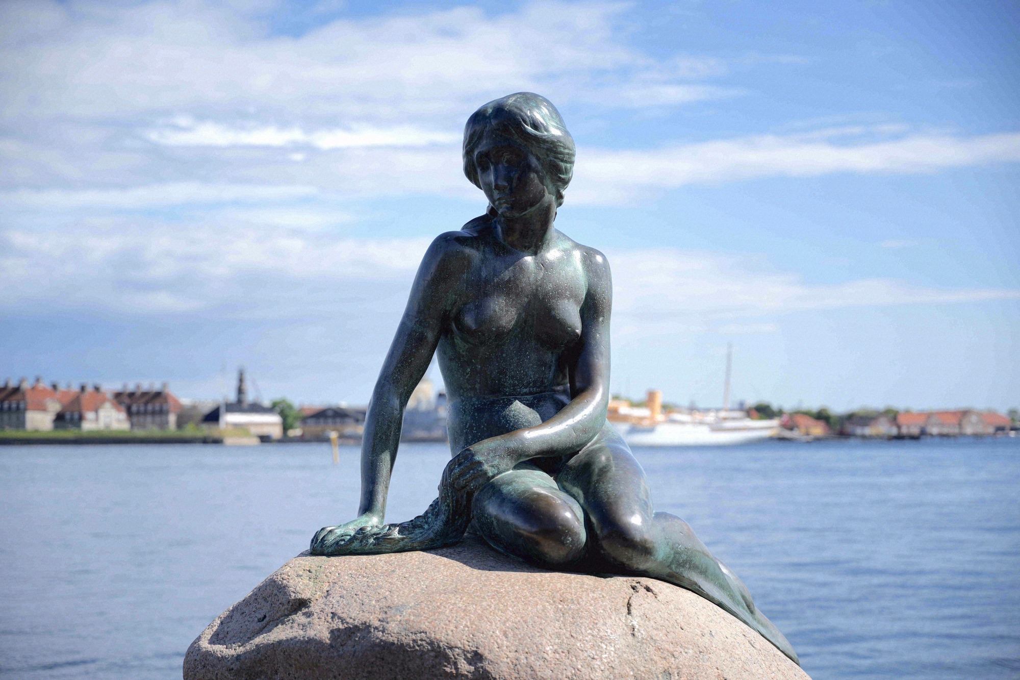 Die beruehmte Bronzestatue der Kleinen Meerjungfrau sitzt auf einem Felsen am Kopenhagener Hafen mit dem Stadtpanorama im Hintergrund.
