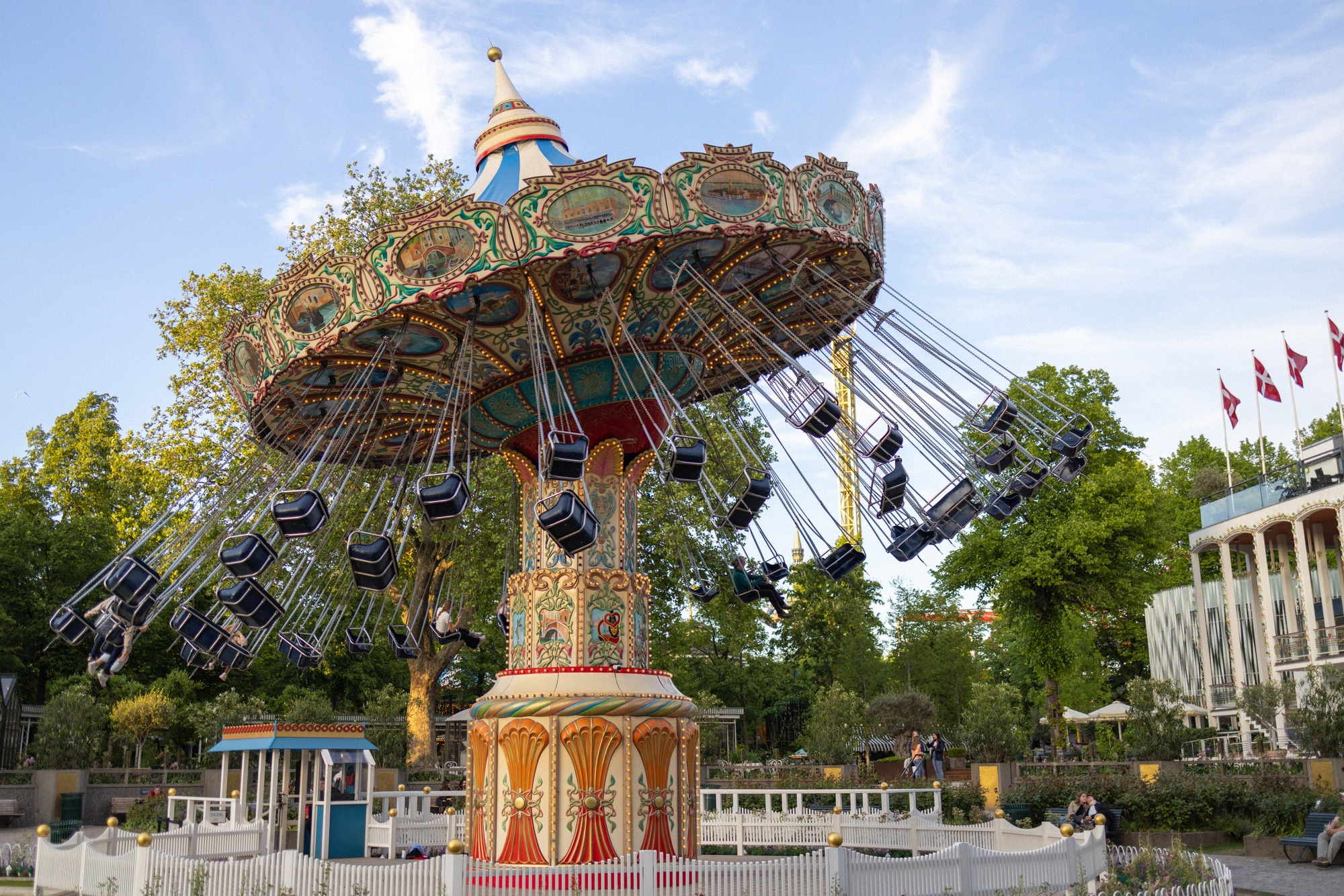 Das bunte Kettenkarussell im Tivoli Gardens dreht sich vor blauem Himmel, im Hintergrund dänische Flaggen und historische Parkgebaeude.